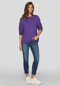 Lila gestrickter Pullover mit einem strukturierten Muster und weitem Ausschnitt, kombiniert mit eng anliegenden blauen Jeans und weißen Sneakers.