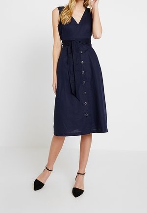 Donna che indossa un abito midi blu navy senza maniche con scollo a V, bottoni frontali e cintura annodata in vita, abbinato a scarpe nere con cinturino alla caviglia.