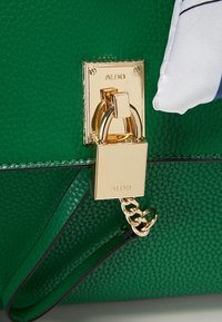 Sac en cuir vert texturé avec serrure et chaîne dorées, partiellement recouvert de tissu blanc à rayures bleues.