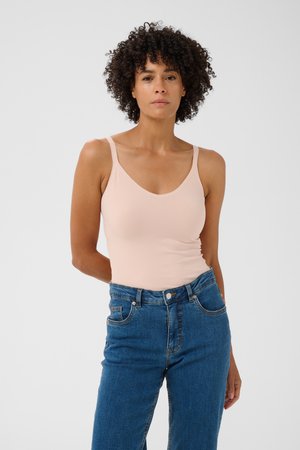 Femme aux cheveux bouclés portant un haut sans manches rose clair et un jean bleu, debout devant un fond clair uni.