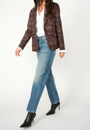 Woman wearing a brown floral blazer, white blouse with frilled cuffs, blue jeans, and black pointed high-heeled ankle boots, walking.