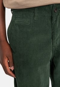 Green corduroy pants with vertical ribbed texture, featuring a back pocket and a front button closure. Material appears soft and dense.