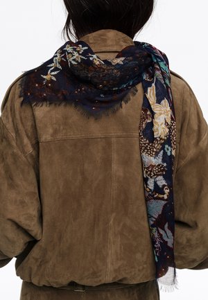 Osoba ubrana w brązową zamszową kurtkę z ciemnym szalem w kwiatowy wzór zarzuconym na ramię i zwisającym z tyłu.