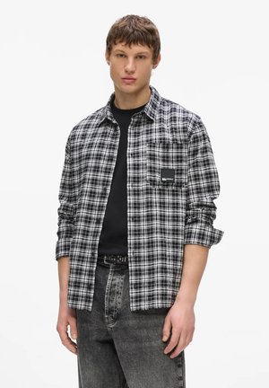 Młody mężczyzna ubrany w czarno-białą koszulę w kratę na czarnym t-shircie z podwiniętymi rękawami i wyblakłych czarnych dżinsach na białym tle.