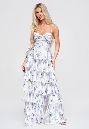 Femme blonde portant une robe blanche à volants superposés, imprimé floral violet et bretelles spaghetti, debout devant un fond clair.