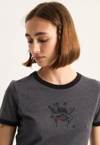 Grå t-shirt med sort krave og ærmeakcenter, prydet med et trykt grafisk design i sort og rød, der viser et skellet med vinger og stjerner.