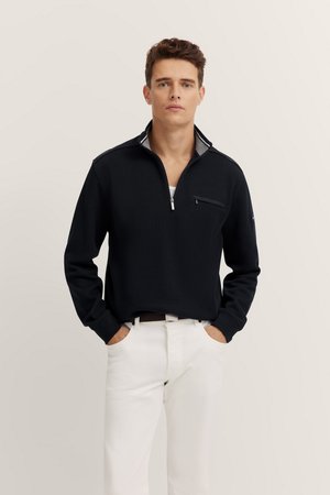 Schwarzer Pullover mit einem Viertelreißverschluss, kontrastierender grauer Innenfutter und einer Brusttasche. Hat lange Ärmel und eine lockere Passform, kombiniert mit weißen Hosen.