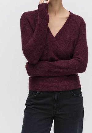 Personne portant un pull à col en V duveteux violet foncé à manches longues, associé à un jean noir taille haute, sur un fond uni.