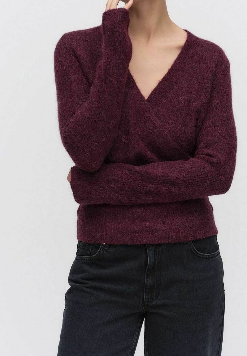 Personne portant un pull à col en V duveteux violet foncé à manches longues, associé à un jean noir taille haute, sur un fond uni.