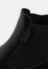 Bottine noire avec une tige en cuir lisse et des panneaux élastiques texturés. Comprend un logo subtil sur le côté.