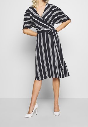 Kobieta w czarnej sukience kopertowej z pionowymi i ukośnymi białymi paskami, z paskiem w talii i białych szpilkach, stojąca z rękami na biodrach.