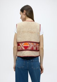 Μπεζ γιλέκο από φλις με πολύχρωμη γεωμετρική κεντητική στη μέση. Φινιρισμένο με κόκκιν ο τελείωμα, φορεμένο πάνω από λευκό πουκάμισο και μπλε τζιν.