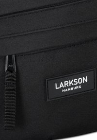 Mochila de tela negra con una textura suave, un cierre de cremallera y una etiqueta de marca que dice "LARKSON HAMBURG" en letras blancas.