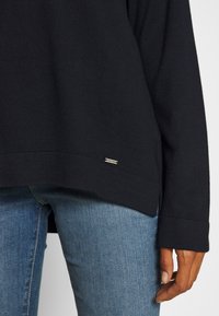 Personne portant un haut noir à manches longues et un jean bleu, avec une main détendue le long du corps.