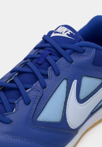 Pogled od blizu na plavi Nike športni copat s vezalkami, mrežastimi paneli, belim swoosh logotipom in označenim jezikom na beli podlagi.