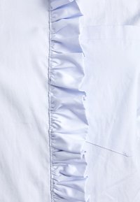 Ljusblå skjorta med volangdetalj framtill och en liten ficka. Tyget verkar slätt med en något glänsande yta.