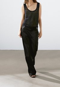 Czarny top na ramiączkach z okrągłym dekoltem, zestawiony z luźnymi czarnymi spodniami z kwadratowymi akcentami i gładką fakturą. Proste sandały.