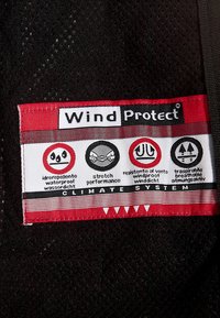 WindProtect-Label, das folgende Eigenschaften zeigt: wasserdicht, dehnbar, winddicht, atmungsaktiv, auf schwarzem strukturiertem Stoff.