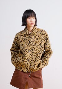 Kabát s leopardím potiskem vyrobený z měkké, texturované látky, s knoflíkovou légou a límcem. Modelka ho kombinuje s hnědou kordovou sukní.