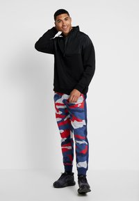 Pullover nero con colletto a zip mezza, abbinato a pantaloni jogger in camouflage multicolore nei toni del rosso, blu e bianco, e scarpe da ginnastica nere.