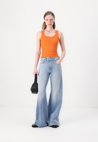 Top de tirantes naranja con relieve, jeans de pierna ancha azul claro, bolso negro sostenido en la mano derecha, modelo de pie contra un fondo liso.