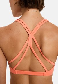 Korallenfarbener Sport-BH mit einem Design mit gekreuztem Rücken. Hergestellt aus elastischem Material, mit glatter Textur und verstellbaren Trägern für zusätzliche Unterstützung.