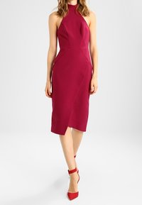 Karmesinrote Neckholderkleid mit tailliertem Oberteil, geraden Nähten und einem knielangen Rock mit asymmetrischem Saum. Kombiniert mit roten Riemchen-Absätzen.