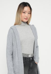Mlada žena s dolgimi ombre lasmi, oblečena v sivi pulover, svetlo sivi cardigan in temno sivo krepkih hlačah z visokim pasom, z rokama v žepih.