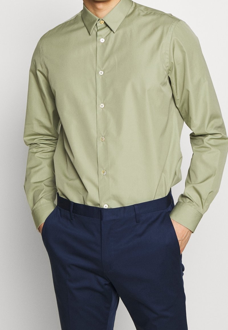 Camicetta verde chiaro a maniche lunghe con colletto a punta e bottoni in colori contrastanti, abbinata a pantaloni blu navy.