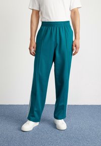 Teal sportieve broek met een elastische tailleband, witte zijstrepen en een losse pasvorm. Gepaarde met witte sneakers op een blauwe vloer.