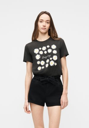 Junge Frau trägt ein schwarzes T-Shirt mit weißen Gänseblümchen und dem Schriftzug "Daisy days", kombiniert mit schwarzen, hoch taillierten Shorts mit einer vorderen Schnürung als Gürtel.