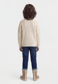 Kind mit lockigem braunem Haar, das einen cremefarbenen Strickpullover, hochgekrempelte dunkle Jeans und hellbraune Gummistiefel trägt, stehend mit dem Rücken zur Ansicht vor einem einfarbigen Hintergrund.