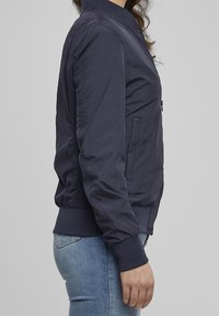Personne portant une veste bomber bleu marine avec des poignets côtelés et un jean bleu clair, debout de profil devant un fond gris uni.