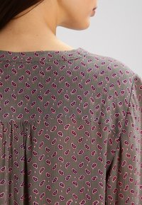 Blouse multicolore en tissu vert olive clair avec petits motifs floraux roses et violets, détail plissé au dos, encolure ronde.