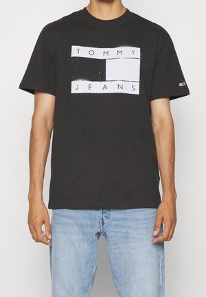 Mężczyzna ubrany w czarną koszulkę z krótkim rękawem z białym logo "Tommy Jeans" oraz jasnoniebieskie jeansy, stojący na jednolitym tle.