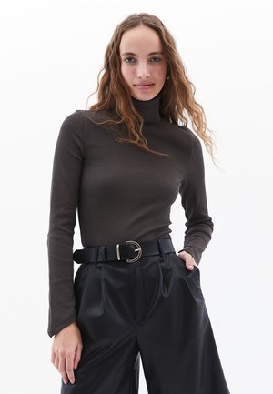 Brauner gerippter Rollkragenpullover mit langen Ärmeln, kombiniert mit hochgeschnittenen schwarzen Hosen, die mit einem Gürtel mit goldener Schnalle verziert sind.