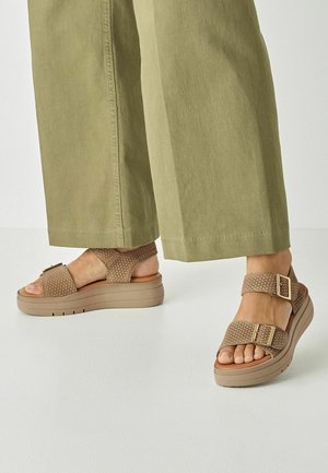 Fødder iført beige vævede plateau-sandaler med guldlåse og olivengrønne bukser med vide ben mod en hvid baggrund.