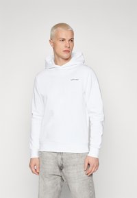 Bílá mikina s kapucí vyrobená z jemné tkaniny, s malým černým logem Calvin Klein na hrudi a žebrovanými manžetami a spodním lemem.