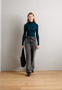 Svarte og grå flared jeans kombinert med en tettsittende turkis ribbe med høy hals, og en svart tote bag med vertikale striper. Treverk gulv, enkel vegg.