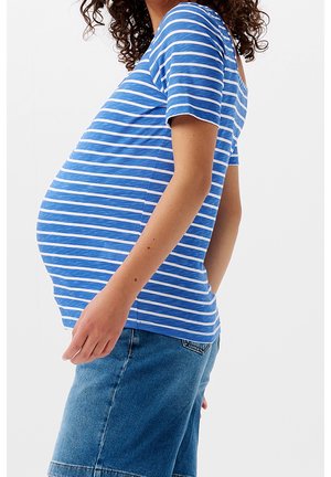 Gravid person iført en blå- og hvidstribet skjorte og jeansnederdel, stående i profil mod en ensfarvet baggrund.