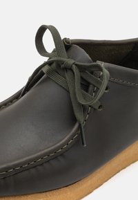 Mörkbrun lädersko med en slät yta, olivgröna snören och kontrasterande sömmar. Har en beige gummisula.