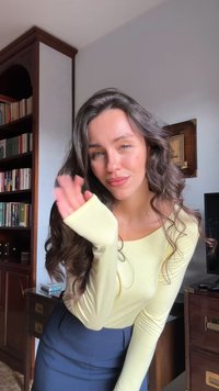 Mlada ženska z dolgimi valovitimi lasmi, oblečena v rumeno majico in modro krilo, maha proti kameri v notranjosti ob lesenem pohištvu in policah za knjige.