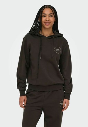 Giovane donna con capelli intrecciati che indossa una felpa con cappuccio marrone scuro abbinata e pantaloni della tuta con piccoli loghi circolari bianchi.