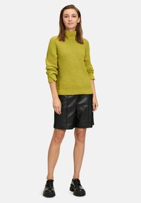 Strick-Turtleneck-Pullover in Limettengrün, mit einem strukturierten Muster und umgeschlagenen Bündchen, kombiniert mit schwarzen Leder-Shorts und klobigen schwarzen Schuhen.