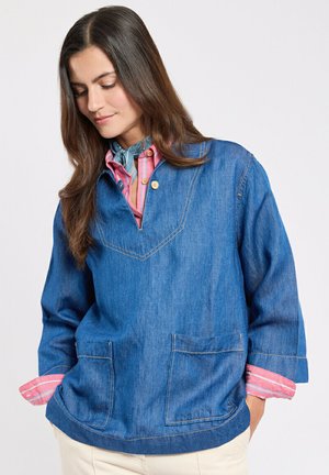 Femme aux longs cheveux bruns portant un pull en denim bleu superposé sur une chemise à rayures roses, mains dans les poches, regardant vers le bas, fond neutre.