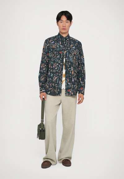 Camicia a maniche corte con stampa grafica colorata su sfondo scuro, abbinata a pantaloni beige chiaro e mocassini marroni, mentre tiene una borsa verde.