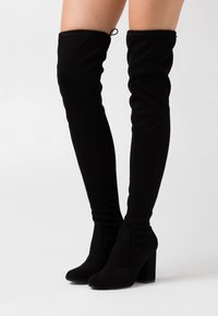 Bottes noires montantes au-dessus du genou en tissu doux, avec un design simple, un bout pointu et un talon épais pour plus de stabilité. Aucun élément métallique visible.