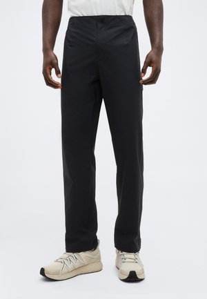 Mann trägt schwarze gerade geschnittene Hose und beige Sportschuhe, steht vor einem schlichten hellen Hintergrund.