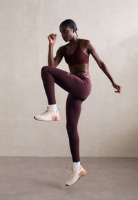 Soutien-gorge de sport bordeaux, leggings taille haute assortis avec logo Nike. Baskets neutres avec des accents roses et oranges. Modèle dans une pose active.