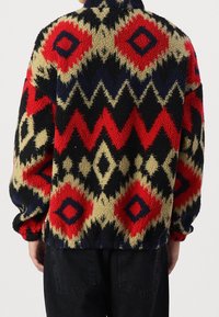 Fuzzy fleecejakke med et flerfarvet geometrisk mønster i rød, sort, creme og marineblå. Høj krave og elastiske mansjetter.